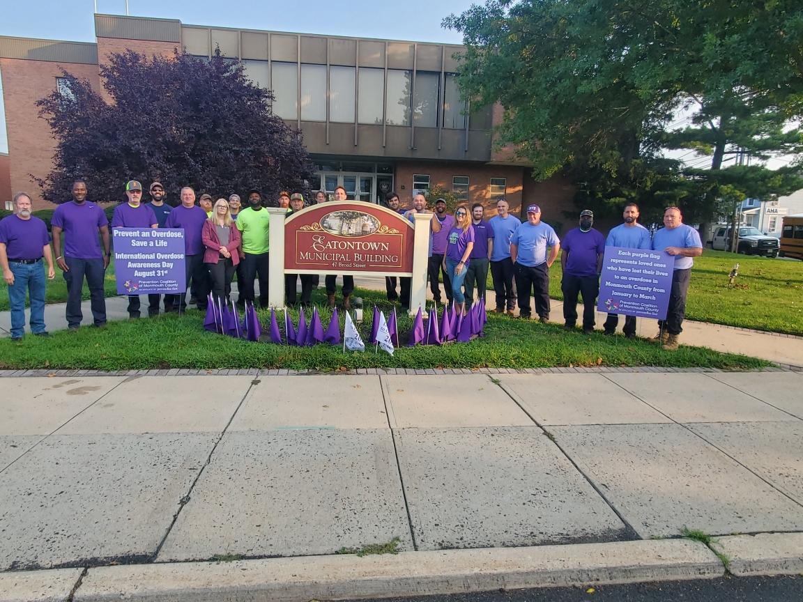 Volunteers stand outside an apartment building sign, holding purple bags for an overdose prevention awareness event.
